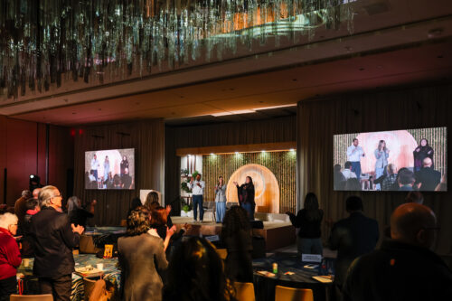 Attendees give a standing ovation during a conference panel discussion, with speakers on stage, presentation screens, and modern event lighting in a large meeting venue.