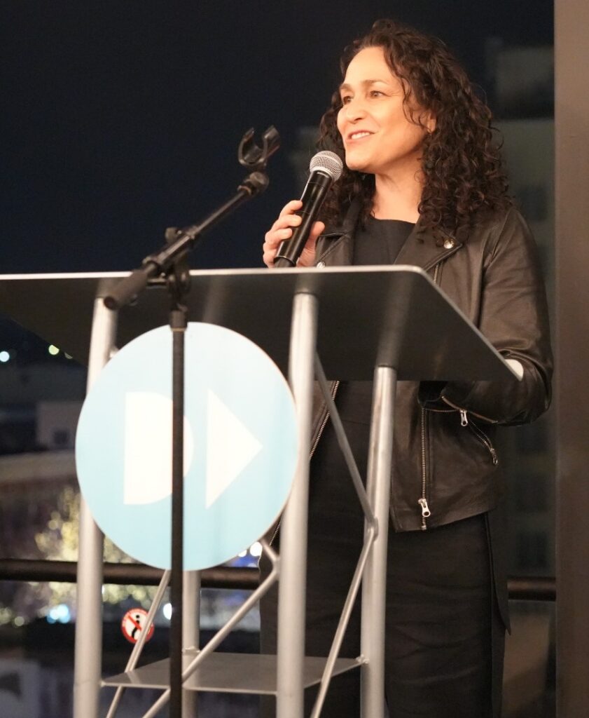 Pamela Shifman addresses attendees from a podium at an evening event, with city lights visible through floor-to-ceiling windows in the background.