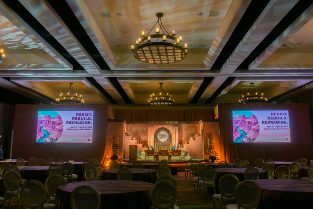 Conference venue with a professionally designed stage, audience seating, and presentation screens displaying Democracy Alliance event branding in Albuquerque, New Mexico.